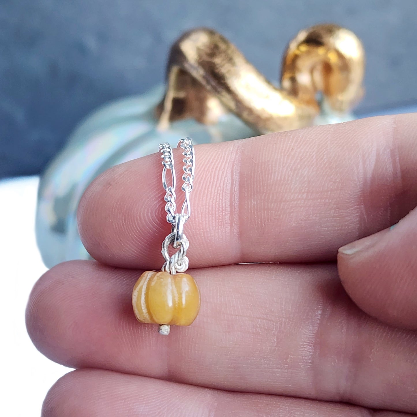 Fingers holding small orange gemstone pumpkin necklace, pumpkin in background